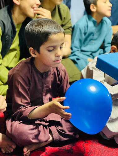 A little boy setting, and holding a blue balloon in his hands.